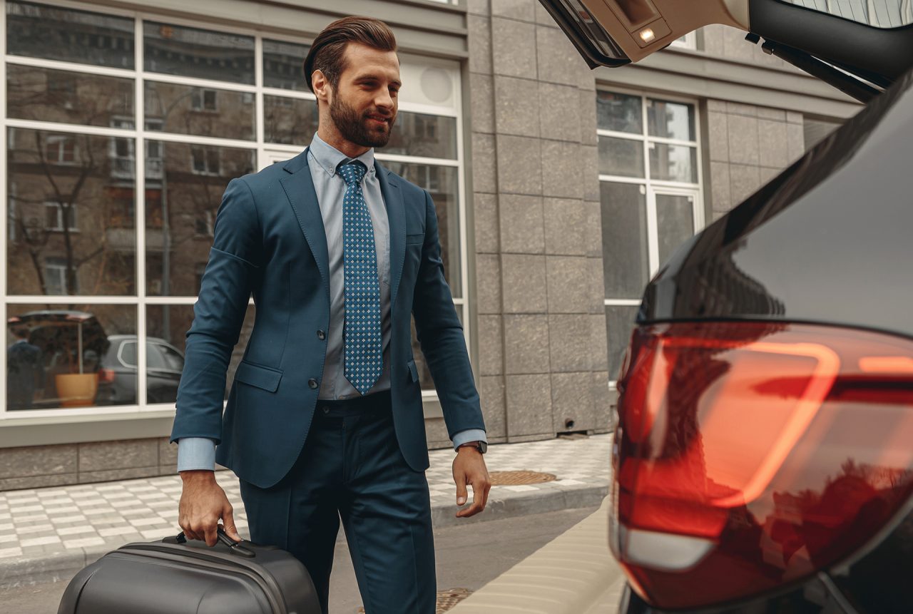 Waist up portrait of handsome Caucasian man with suitcase on wheels in hand standing near the car in the outdoors